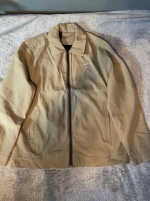 GANT Harrington Jacka Beige L (passar M) | Old Money Preppy Minimalist Clean Fit - Stilren och tidlös GANT Harrington jacka i beige med minimalistisk design och broderad logotyp på bröstet. Perfekt för en clean old money / preppy look och funkar lika bra till vardag som till mer uppklädda outfits. Lätt att matcha med jeans, chinos eller cargos.  Lätt och bekväm passform med dragkedja fram och diskreta fickor för ett clean uttryck. Perfekt vår-, sommar- och höstjacka 🤍 • Märke: GANT • Storlek: L (passar även M beroende på fit) • Skick: Mycket fint • Unisex • Snabb leverans