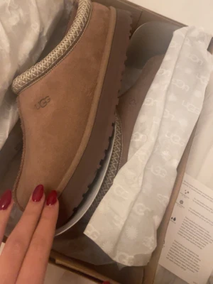 Bruna UGG tofflor i mocka - Säljer ett par bruna UGG tofflor i mjuk mocka med dekorativt mönster runt öppningen. Tofflorna har en rejäl sula och klassisk UGG-logga på sidan. Perfekta för att hålla fötterna varma och sköna hemma. Ett litet litet hål på sulan typ 