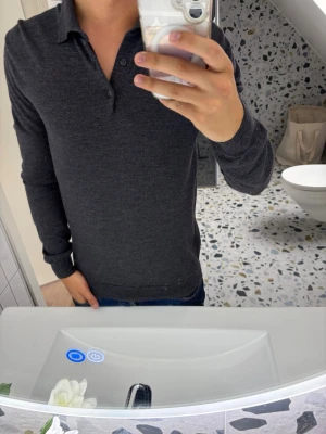 Mörkgrå långärmad pike från J Lindeberg  - Snygg mörkgrå piké med lång ärm och klassisk krage. Skjortan har knappar vid halsen och är tillverkad i ett mjukt material som känns skönt mot huden. Perfekt till jeans för en clean och enkel look.