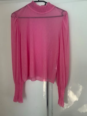 Rosa blus med volangkrage - Trendig rosa blus i transparent material med hög volangkrage och långa puffärmar. Ärmsluten har breda smockdetaljer som ger en snygg siluett. Perfekt för dig som vill sticka ut med färg och stil. Kom gärna med förslag på pris. 