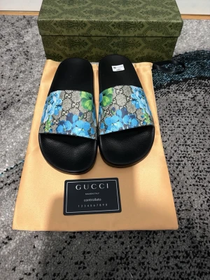 Gucci sandaler med blå blommor - Gucci sandaler med svart sula och bred rem i klassiskt GG-mönster dekorerad med blå blommor och gröna blad. Snygg och unik design som sticker ut, perfekt för dig som vill ha något extra från ett lyxigt märke.