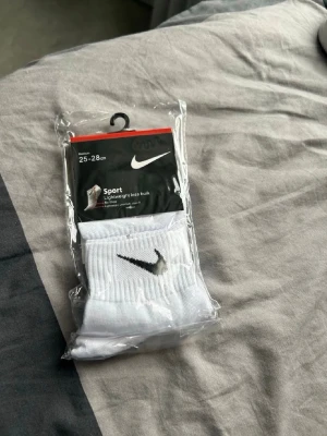 Vita sportstrumpor från Nike - Fräscha vita sportstrumpor från Nike med klassisk svart swoosh-logga. Strumporna är ribbade och tillverkade i ett lätt och bekvämt material som passar perfekt till träning. Storlek 25-28 cm enligt förpackningen.