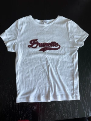 Snygg vit t-shirt med vinröd text - Snygg vit t-shirt från Subdued med vinröd broderad text 'Brunette' på bröstet. Klassisk rund halsringning och korta ärmar. Tillverkad i mjukt bomullsmaterial, perfekt för en avslappnad och trendig look. Storlek är M men är väldigt liten i storleken🤍 bara använd två gånger