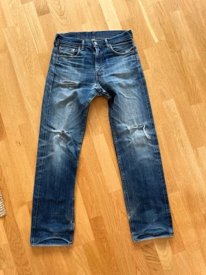 Selvedge Levis jeans - Säljer ett par blå Levi's jeans med raka ben och slitningar på knäna och vid bensluten. Klassisk femficksmodell med coolt sliten look och tydliga tvätteffekter. Jeansen har mycket täcken på användning (se bilder) men går lätt att fixa med fina lagningar. Jeansen har även hål i den högra fickan. Hör av er om ni har frågor!!