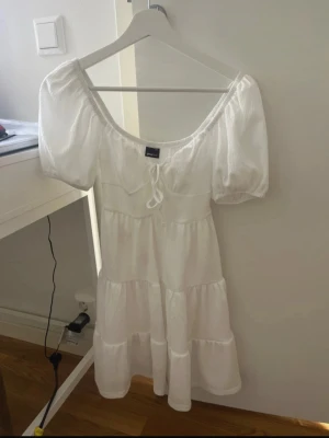 Vit kort klänning med puffärm - Söt vit kort klänning från Gina Tricot med puffärmar och knytning framtill. Klänningen har en luftig och flowy passform med volangdetaljer och är perfekt för varma dagar. Gjord av polyester. Storlek XXS. Finns att hämta i Partille eller fraktas mot att köparen står för frakten🌷