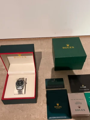 Role klocka med svart urtavla - Snygg Role klocka i silver med svart urtavla och länkarmband i metall. Urtavlan har markerade index och boetten är dekorerad med stenar runt om. Klockan levereras i originalbox med tillhörande certifikat och dokumentation.