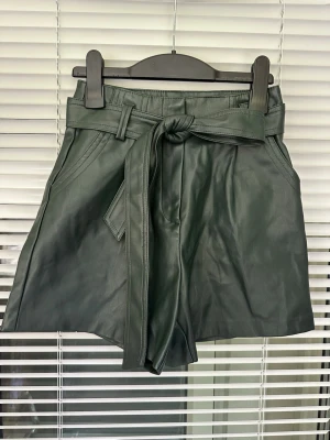 Mörkgröna shorts i skinnimitation Amisu storlek: 32 - Snygga mörkgröna shorts i skinnimitation från Amisu. Högmidjade med bred knytskärp i midjan och fickor på sidorna. Perfekta för dig som vill ha en trendig och edgy look.