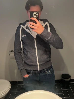Mörkgrå hoodie från Superdry - Mörkgrå hoodie från Superdry med vit dragkedja och vita snören i huvan. Klassisk modell med kängurufickor framtill och ribbade muddar vid ärmslut och nederkant. !Defekt finns på sista bild! Inget stort går nog och limma 