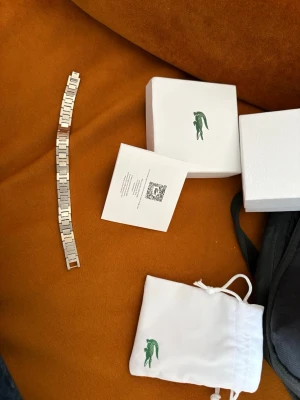 Lacoste silverfärgad klocklänk - Snygg silverfärgad klocklänk från Lacoste i rostfritt stål. Länken har en stilren och modern design med rektangulära länkar och passar perfekt till din Lacoste-klocka. Levereras med originalförpackning och tygpåse med den klassiska gröna krokodilloggan.