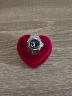 Silverfärgad ringklocka  - Unik silverfärgad ring med en liten svart urtavla och vita siffror. Ringen har en klockdesign och elastiskt band som gör att den passar de flesta fingrar. Perfekt accessoar för dig som gillar ovanliga smycken.