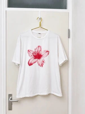 Vit t-shirt med rosa blomma - Vit t-shirt med en stor rosa blomma tryckt på framsidan. T-shirten har rund halsringning och korta ärmar. Perfekt för en avslappnad och trendig stil.
