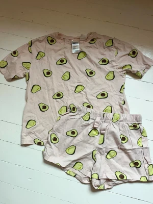 Rosa pyjamas med avokadotryck - Säljer ett set med pyjamas i mjuk bomull, bestående av en rosa t-shirt och matchande shorts. Båda delarna har ett gulligt avokadotryck över hela plagget. Perfekt för mysiga hemmakvällar eller chill dagar. T-shirt och shorts med resår i midjan. Använder inte längre så säljer.
