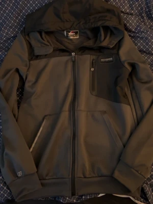 Grå/Svart zip hoodie med huva från McKenzie - Snygg svart vindjacka från McKenzie med huva och dragkedja framtill. Jackan har en bröstficka med dragkedja, två sidofickor och diskreta logodetaljer. Perfekt för blåsiga dagar och enkel att matcha med olika outfits. Pris kan diskuteras