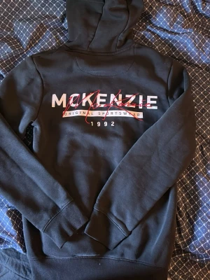 Svart hoodie från McKenzie - Svart hoodie från McKenzie med stor logga och textdetaljer i vitt och rött på ryggen samt mindre logga på bröstet. Klassisk huva med dragsnören och magficka framtill. Tillverkad i mjukt bomullsmaterial, perfekt för en avslappnad streetwear-look. Pris kan diskuteras 