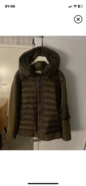 Olivgrön pufferjacka från Moncler - Snygg olivgrön pufferjacka från Moncler med stor huva och quiltad front. Jackan har stickade ärmar och detaljer i nylon, samt en ficka på ärmen med Moncler-logga. Perfekt för kalla vinterdagar och riktigt trendig look.