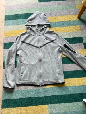 Grå orginel Nike Tech hoodie - Snygg grå hoodie från Nike med dragkedja framtill och svart Nike-logga på bröstet. Hoodien har svarta detaljer över bröstet och en ficka med dragkedja på ena ärmen. Tillverkad i mjukt material och har två sidofickor. Perfekt för chill eller träning.