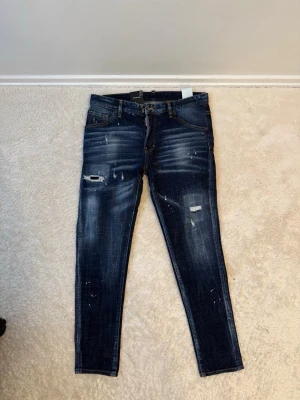 Mörkblå distressed slim fit jeans - Snygga mörkblå slim fit jeans från DSQ med slitningar, blekta partier och färgstänk för en trendig, avslappnad look. Klassisk femficksmodell med dragkedja och knapp. Perfekta för dig som gillar streetstyle och vill sticka ut.