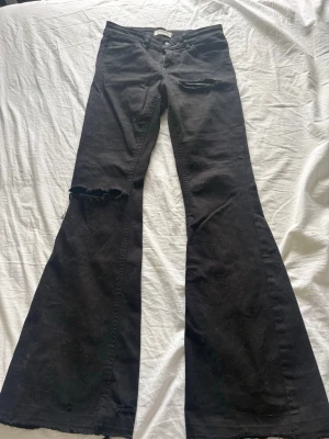 Svarta utsvängda jeans med slitningar - Säljer ett par svarta jeans med utsvängda ben och slitna detaljer vid knä och ficka. Jeansen har råa kanter nertill och en cool, trendig look. Perfekta för dig som gillar en avslappnad och edgy stil.
