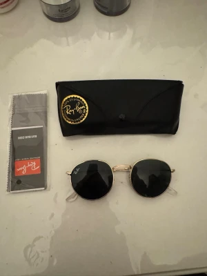 Ray-Ban runda solglasögon guld - Snygga Ray-Ban solglasögon med rund form och tunna guldfärgade metallbågar. Svarta glas och klassisk Ray-Ban-logga på linsen. Kommer med originalfodral och putsduk. Perfekta för dig som vill ha en tidlös och trendig look.
