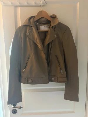Olivgrön skinnjacka från Selected Femme - Snygg grå/beige skinnjacka från Selected Femme med asymmetrisk dragkedja, bred krage och två snedställda fickor med dragkedjor. Jackan har detaljer vid midjan och dragkedjor vid ärmsluten för extra edge.