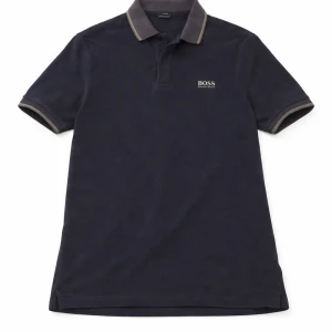 Mörkblå piké från BOSS - Klassisk mörkblå pikétröja från BOSS med korta ärmar och kontrastfärgade detaljer på krage och ärmslut. Tröjan har knappar vid halsen och diskret BOSS-logga på bröstet. Perfekt för en clean och stilren look.