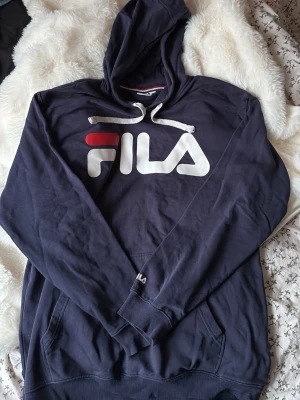 Mörkblå hoodie från FILA - Snygg mörkblå hoodie från FILA med stor logga i vitt och rött på bröstet och mindre logga på ärmen. Hoodien har huva med vita dragsnören och en klassisk magficka. Tillverkad i mjukt bomullsmaterial, perfekt för en avslappnad streetwear-look.