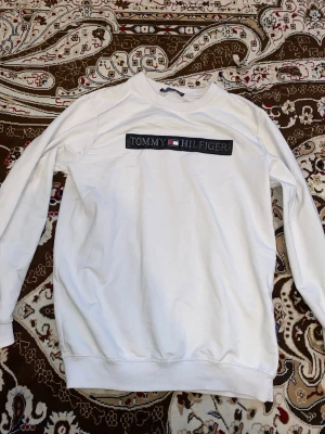 Vit sweatshirt från Tommy Hilfiger - Säljer en vit sweatshirt från Tommy Hilfiger med klassisk logga och flagga broderad på bröstet. Tröjan har rund halsringning, långa ärmar och ribbade muddar vid ärmslut och nederkant. Perfekt för en clean och stilren look.