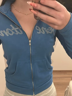 Blå zip hoodie från Benetton - Snygg blå hoodie från Benetton med dragkedja framtill och två stora fickor. Dekorerad med glittrande stenar som stavar Benetton över bröstet. Tillverkad i mjukt material och har en sportig, avslappnad look. Perfekt för dig som vill ha en chill och trendig stil.
