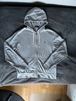 Grå zip hoodie från Polo Ralph Lauren - Snygg grå zip hoodie från Polo Ralph Lauren med vit dragsko i huvan och klassisk blå logga broderad på bröstet. Tillverkad i mjukt bomullsmaterial och har två fickor framtill. Perfekt för en avslappnad och stilren look.