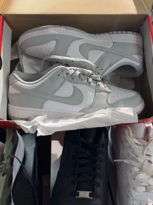 Nike Dunk Low vit/grå sneaker - Säljer ett par Nike Dunk Low sneakers i vitt och ljusgrått. Skorna har klassisk låg siluett, perforerad tåbox och ikoniska Swoosh-loggan på sidan. Tillverkade i slittåligt läder med vit sula och matchande grå snören. Perfekta för dig som gillar clean och stilren look.