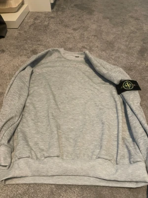 Ljusgrå sweatshirt från Stone Island - Säljer en stilren ljusgrå sweatshirt från Stone Island med klassisk rund halsringning och ribbade muddar. Tröjan har den ikoniska Stone Island-loggan på ärmen och är tillverkad i mjukt bomullsmaterial. Perfekt för en avslappnad look.
