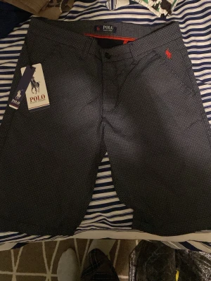 Mörkblå mönstrade shorts från Polo Ralph Lauren - Snygga mörkblå shorts från Polo Ralph Lauren med diskret rutigt mönster och röd logga broderad på sidan. Tillverkade i 100% bomull för en skön känsla. Klassisk passform med knapp och dragkedja framtill.