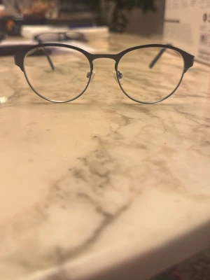 Runda glasögon med metallbåge från D.Arnesen - Ny pris 900kr. Snygga glasögon med tunn, rund metallbåge i svart. Klassisk och stilren design som passar till många olika looks. Näskuddar i plast för extra komfort. Perfekt accessoar för dig som vill ha en tidlös vibe. 