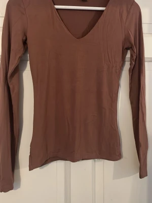Brun långärmad v-ringad topp - Säljer en enkel och stilren brun långärmad topp med v-ringning. Toppen har en slim passform och är tillverkad i ett mjukt och stretchigt material som känns skönt mot huden. Perfekt att matcha med jeans eller kjol för en clean look.