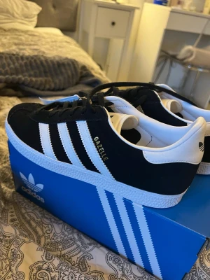 Adidas Gazelle svartvita sneakers - Säljer ett par klassiska Adidas Gazelle sneakers i svart mocka med vita detaljer och de ikoniska tre ränderna på sidan. Skorna har snörning, vit platt sula och guldfärgad Gazelle-text på sidan. Perfekta för dig som gillar retrostil och streetwear. Skorna är aldrig använda då de va för stora. 