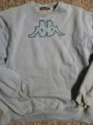 Ljusblå sweatshirt från Kappa - Ljusblå sweatshirt från Kappa med det klassiska broderade logotypmotivet i turkos och lila på bröstet. Tröjan har rund halsringning, ribbade muddar vid ärmslut och nederkant samt en avslappnad passform. Perfekt för en casual streetwear-look. Tyvärr har den några fläckar och har några år på nacken därav priset, men den är klassisk! Storlek XL