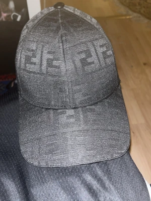 Svart Fendi keps med FF-mönster - Säljer en svart keps från Fendi med det klassiska FF-logomönstret över hela kepsen. Kepsen har böjd skärm och en stilren design som passar till streetwear och modeintresserade. Materialet är tyg med en diskret glans och loggan är invävd i tyget.
