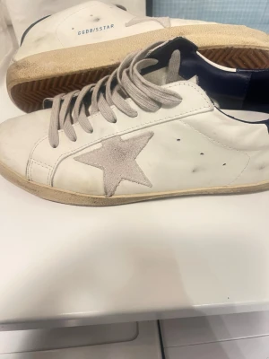Vita Golden Goose sneakers med stjärna - Snygga vita sneakers från Golden Goose med beige mockastjärna på sidan och beige snören. Skorna har mörkblå detalj vid hälen och en vintage-inspirerad sula. Ovandelen är i läder och mocka, med GGDB/SSTAR-tryck på sidan. Perfekta för en trendig streetstyle-look.