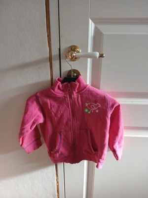 Rosa ziptröja med broderi - Söt rosa ziptröja med hög krage och två fickor framtill. Tröjan har ett litet broderat motiv av en fågel och blommor på bröstet. Tillverkad i mjukt material, perfekt för lager på lager. Långa ärmar och ribbade muddar för extra komfort.