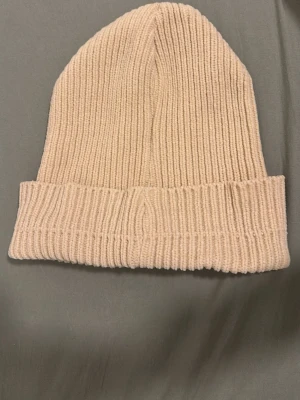 Beige ribbstickad mössa - Säljer en stilren beige ribbstickad mössa med uppvikt kant. Mössan är mjuk och stretchig, perfekt för kyliga dagar. Materialet känns som en blandning av ull och syntet, vilket gör den både varm och bekväm.