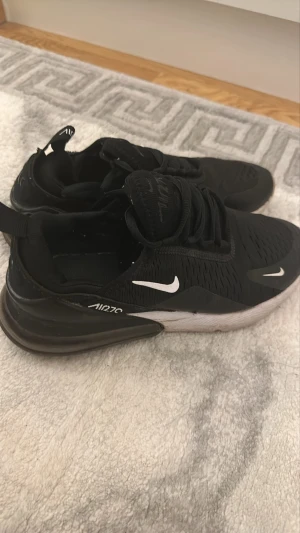 Nike Air Max 270 svarta sneakers - Svarta Nike Air Max 270 sneakers med vit sula och tydlig Air-enhet i hälen. Skorna har mesh-ovandel för bra ventilation, vita swoosh-loggor och en sportig, modern look. Perfekta för dig som gillar streetwear och vill ha bekväma sneakers med snygg design.