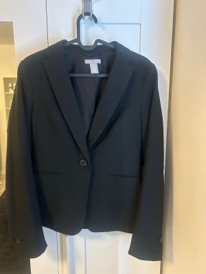 Svart kavaj från H&M, storlek 38 - Klassisk svart kavaj från H&M i storlek 38. Enkelknäppt modell med smal krage och två fickor framtill. Tillverkad i ett slätt tyg med helfodrad insida. Perfekt för en stilren och clean look.