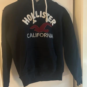 Mörkblå hoodie från Hollister - Mörkblå hoodie från Hollister med vit broderad text och röd logga på bröstet. Tröjan har huva med vita snören, magficka och mjukt grått foder i luvan. Perfekt för dig som gillar en avslappnad och sportig stil.
