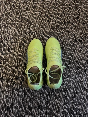 Gula/gröna Puma fotbollsskor med dobbar - Snygga fotbollsskor från Puma i en fräsch gul färg med svarta detaljer och blå sula. Skorna har snörning och är designade för att ge bra grepp på planen med sina fasta dobbar. Perfekta för dig som vill sticka ut på fotbollsplanen.