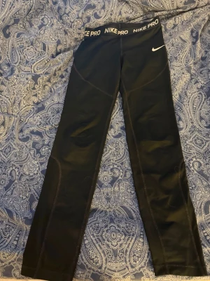Svarta Nike Pro leggings - Säljer ett par svarta Nike Pro leggings med elastisk midja där det står 'NIKE PRO' runtom. De har en tight passform och är tillverkade i ett stretchigt material som passar perfekt till träning eller chill. Swoosh-logga på vänster höft för extra sportig vibe.