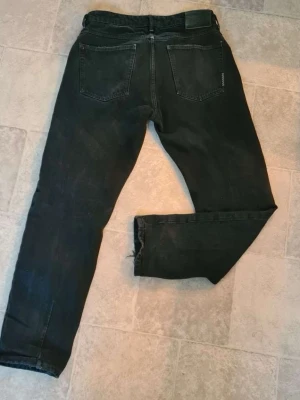New Stockholm jeans  - Säljer ett par svarta New Stockholm jeans  med rak passform och klassisk  femficksmodell med bälteshällor och knappgylf. Jeansen har en enkel och stilren look som passar till det mesta. Materialet är denim i bomull.
