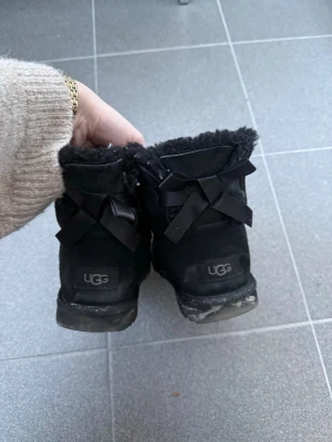 Svarta UGG boots med rosetter - Ge gärna bud eller förlag på pris så kommer vi överens om något bra!!!   Säljer ett par svarta UGG boots i mocka med fluffigt foder och snygga rosetter bak. Klassisk modell som är både varm och trendig, perfekt för kyliga dagar.