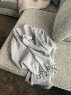 Ljusgrå halsduk med fransar - En stilren ljusgrå halsduk i mjukt material med klassiska fransar i ändarna. Perfekt att svepa runt halsen för extra värme och en clean look. Passar till de flesta outfits och är enkel att matcha med andra färger.