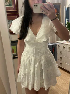 vit studentklänning  - Supersöt vit kortklänning med djup v-ringning, volangdetaljer vid axlar och hals samt broderade blommor över hela tyget. perfekt till studenten 