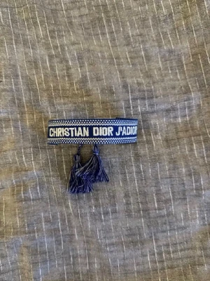 Christian dior armband  - Snyggt blått tygarmband från Christian Dior med texten 'J'ADIOR' broderad i vitt. Armbandet har dekorativa tofsar i ändarna och justerbar knytning. Perfekt accessoar för dig som gillar exklusiva märken och vill addera en trendig detalj till din stil.